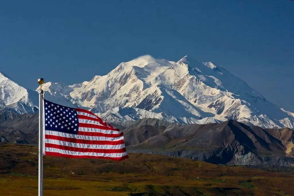 American flag waves in front of snow-capped Denali mountain under a clear blue sky.