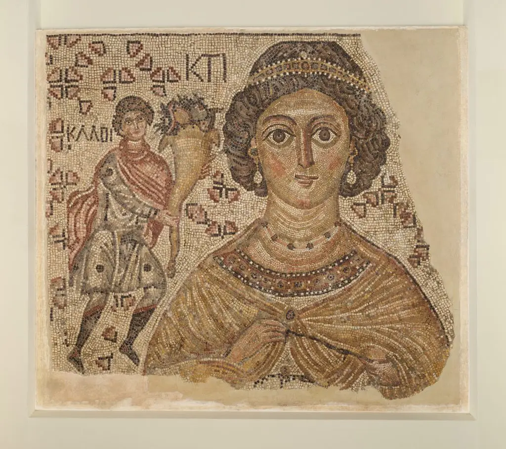 Ancient Byzantine mosaic of a richly dressed woman with a servant, featuring detailed patterns and Greek inscriptions.