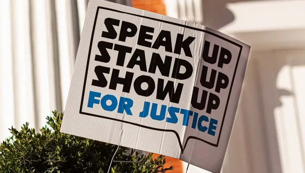 Protest sign reading Speak Up, Stand Up, Show Up For Justice with surrounding greenery and soft background.