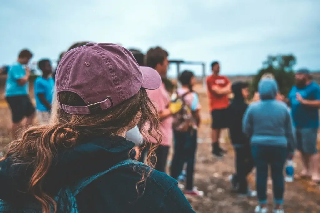 Group of people in casual clothing gathered outdoors, with a person in a purple cap in the foreground.