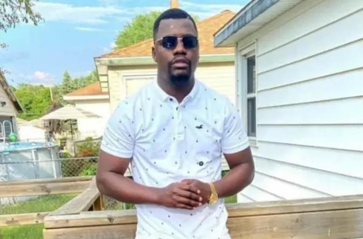 Man in a white shirt and sunglasses standing on a wooden deck in a suburban backyard on a sunny day.