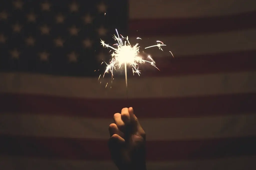 Hand holding a bright sparkler in front of the American flag, symbolizing celebration and patriotism.