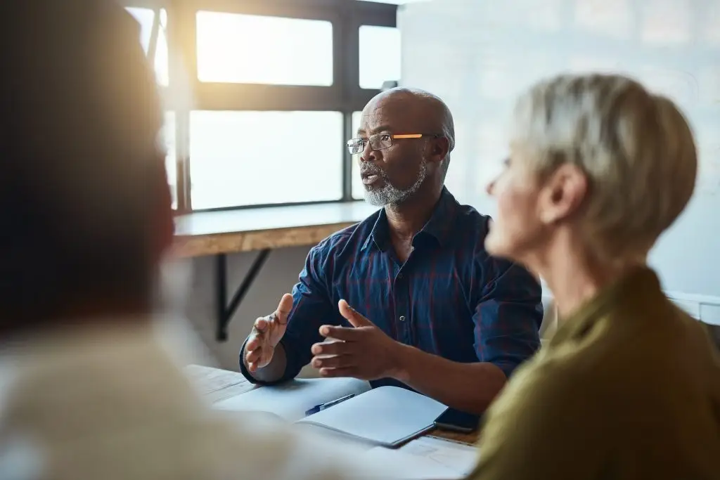 Man in casual meeting, discussing ideas with colleagues around a table in a sunlit office.