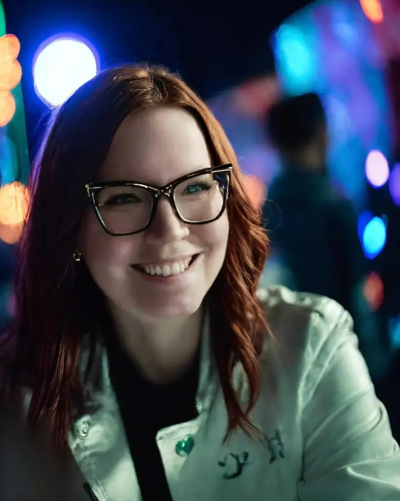 Smiling woman with glasses at a nighttime event with colorful bokeh lights in the background.