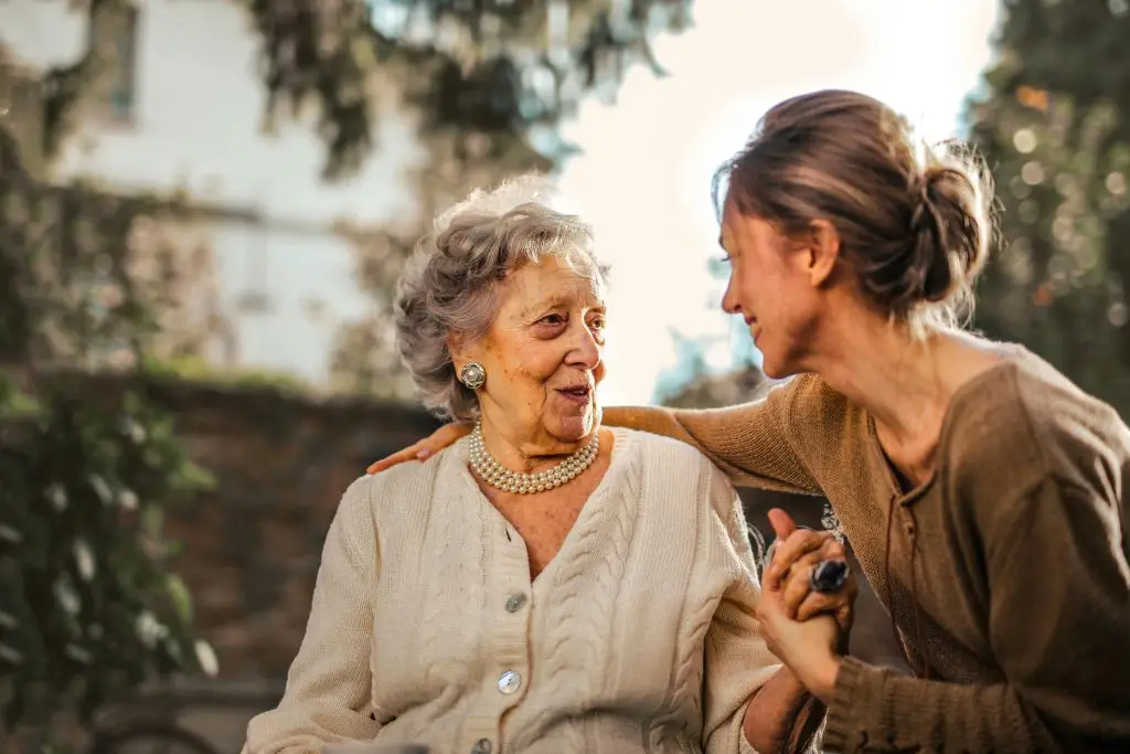 Elderly woman and young woman smiling and holding hands in a park, symbolizing love and companionship.