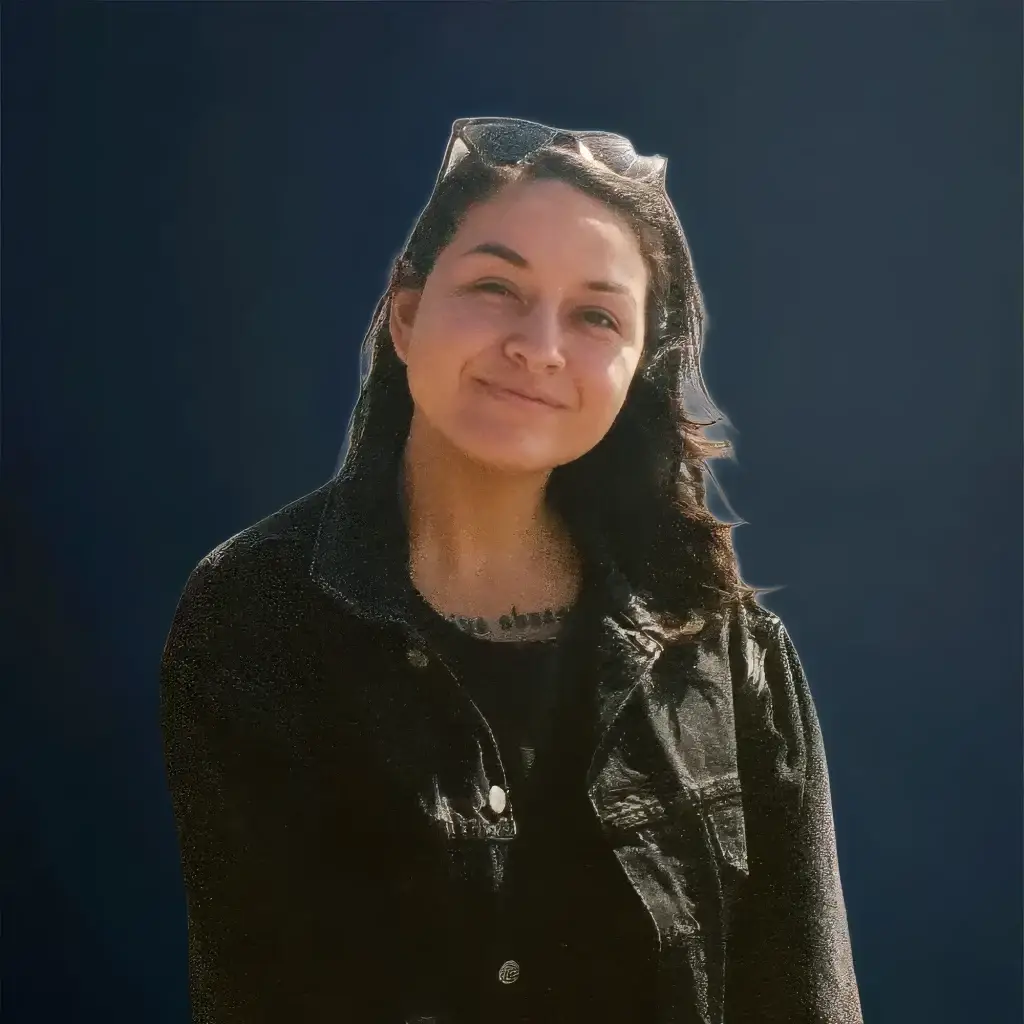 Smiling individual wearing a black jacket and glasses against a dark blue background.