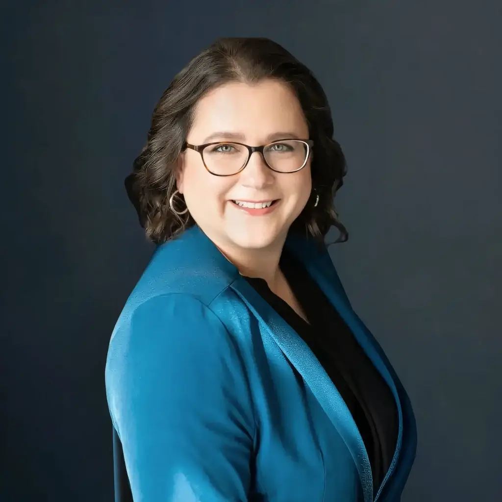 Smiling person in glasses and blue blazer against dark background.