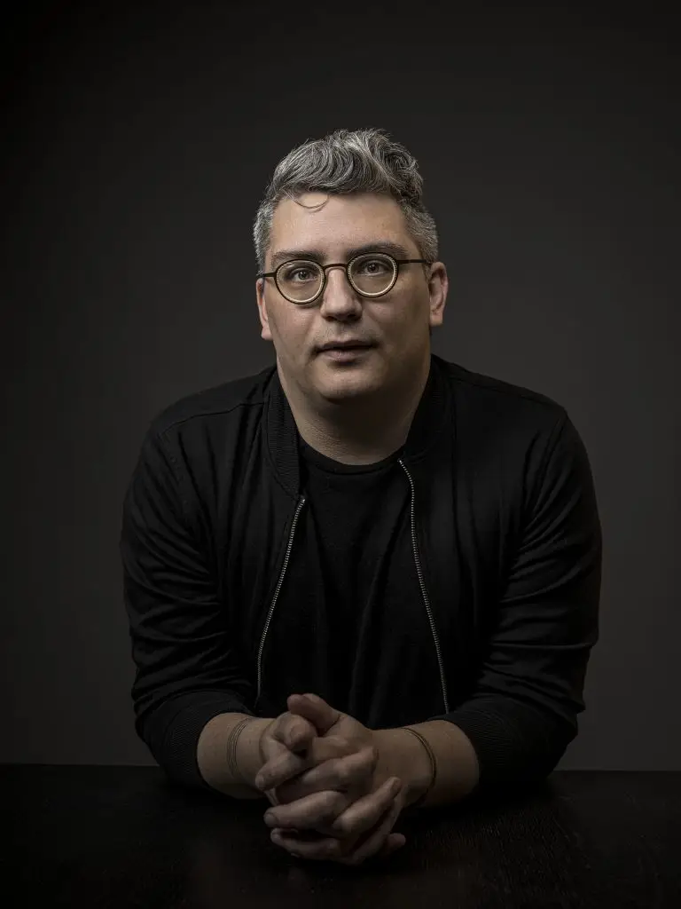 Portrait of a person with glasses, clasped hands, in a dark outfit, against a dark background.