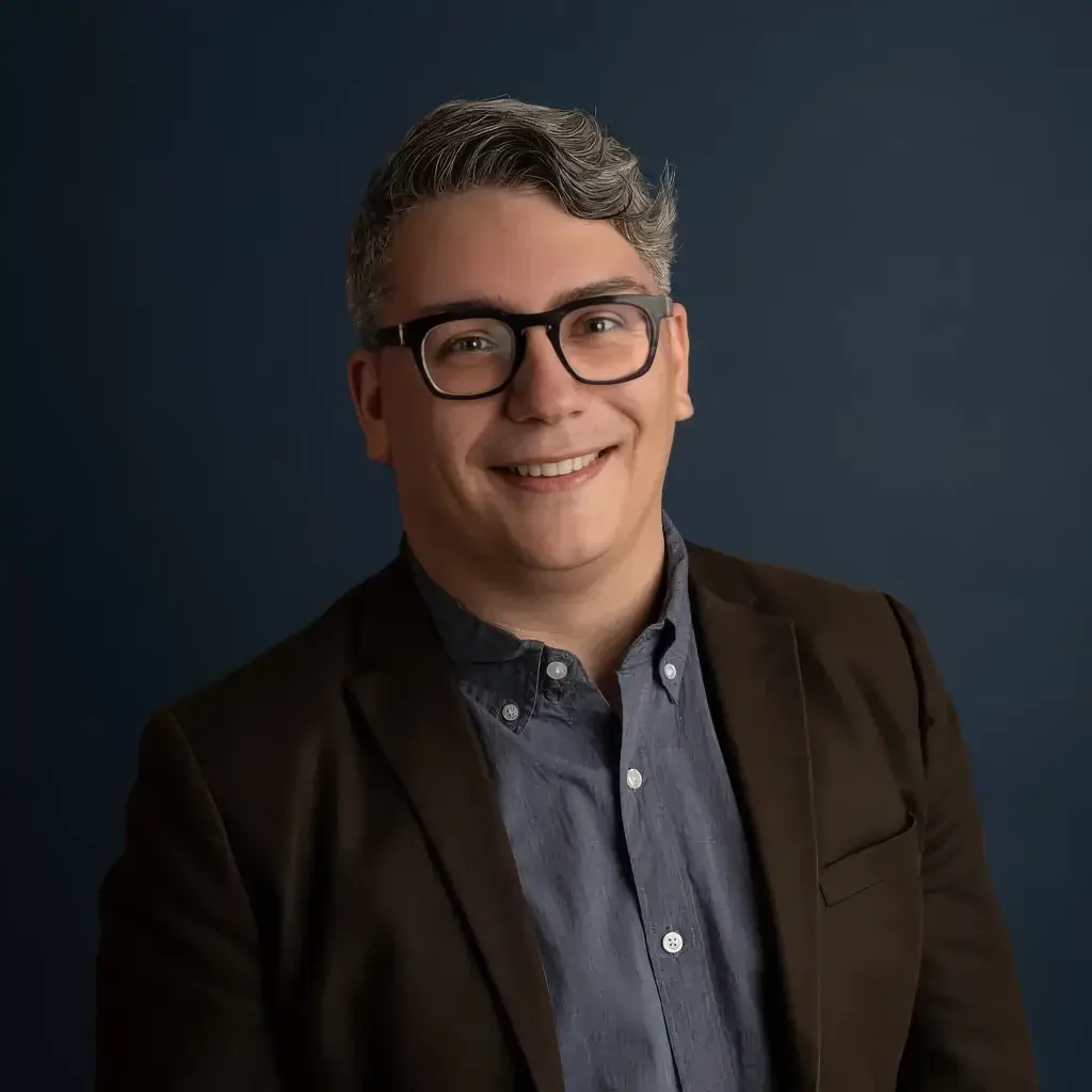 Smiling person in glasses and blazer against a dark background, professional headshot.