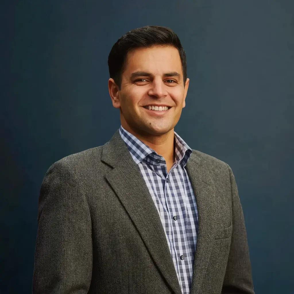 Smiling man in gray blazer and checkered shirt against a dark blue background. Formal portrait photograph.