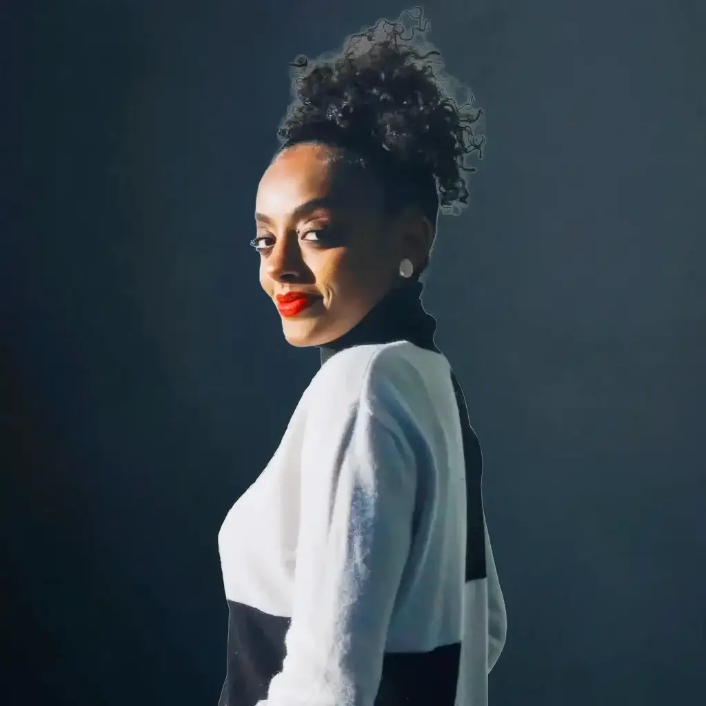 Smiling woman with curly hair, wearing a black and white outfit against a dark background.
