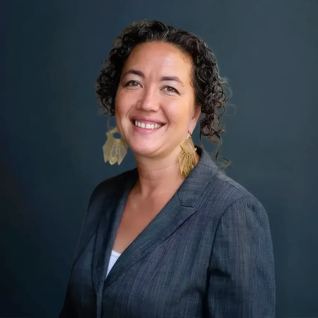 Smiling woman with curly hair, wearing a gray blazer and leaf-shaped earrings, posed against a dark background.