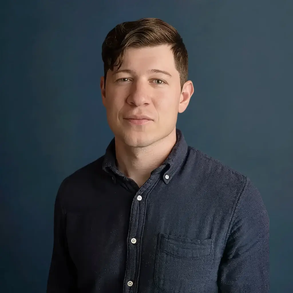 Man in a dark shirt posing against a plain dark background, gazing confidently at the camera.